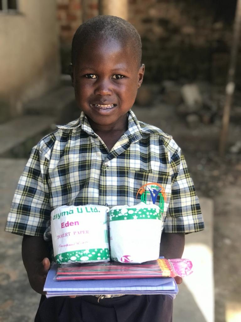 A smiling boy in his school uniform holding exercise books, pencils and toilet rolls for the new term