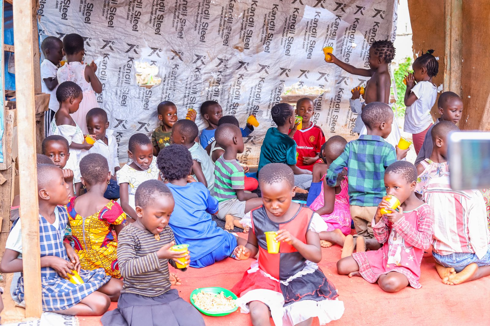 Children sitting together under a shelter, eating and drinking at an outreach day
